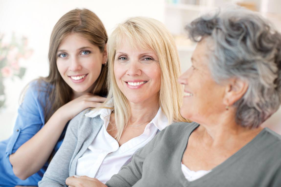 ©istock.com/svetikd Drei Frauen unterschiedlichen Alters sitzen dicht beieinander und lächeln. Die Jüngste hat langes braunes Haar, die Frau mittleren Alters ist blond, und die Älteste hat kurzes graues Haar. Sie wirken glücklich und fühlen sich wohl miteinander.